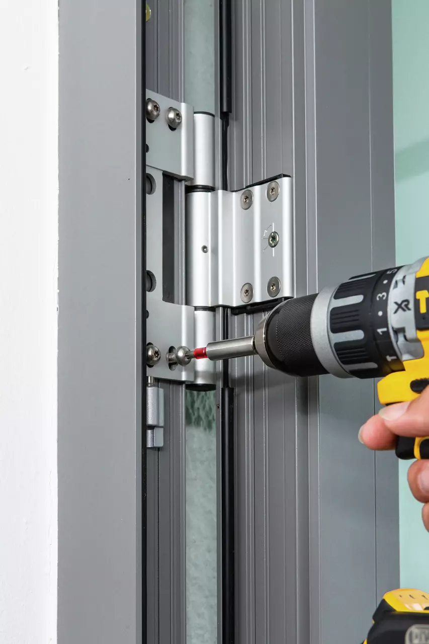 Person using a yellow and black drill to adjust screws on a metal door hinge.
