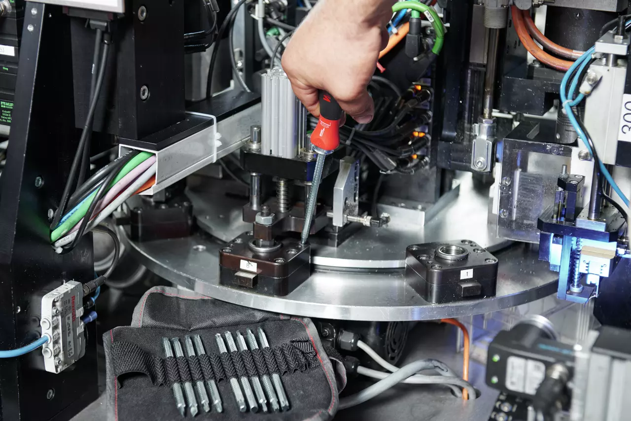 A person uses a red-handled screwdriver on a complex machine, with a set of bits in a pouch nearby.