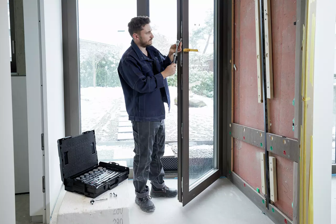 A man working with a wrench on a window frame, toolbox beside him.