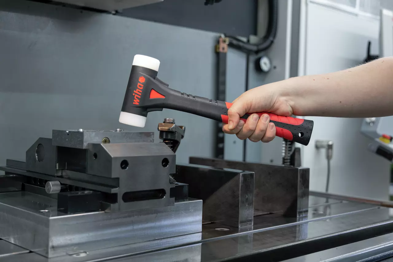 Eine Person hält einen schwarz-roten Schonhammer mit weißen Enden in der Nähe eines Metallschraubstocks.