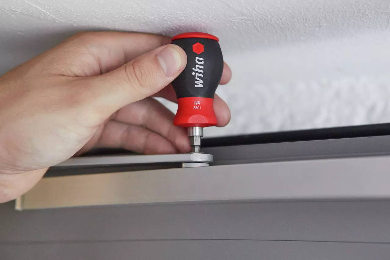 Hand using a red and black screwdriver to adjust a metal fitting on a door frame.