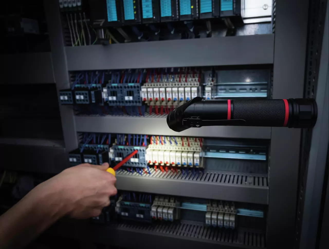A hand holding a screwdriver points at components in an electrical panel, illuminated by a flashlight.