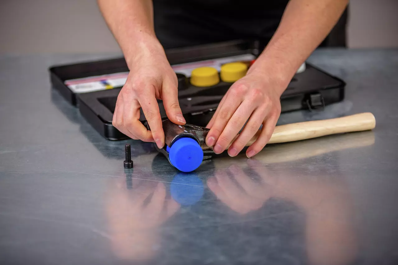 Person assembling a mallet with blue head and wooden handle on a metal surface