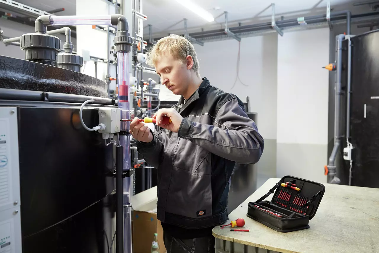 A person using a screwdriver on a pipe system in an industrial setting. A tool case is open on the table.