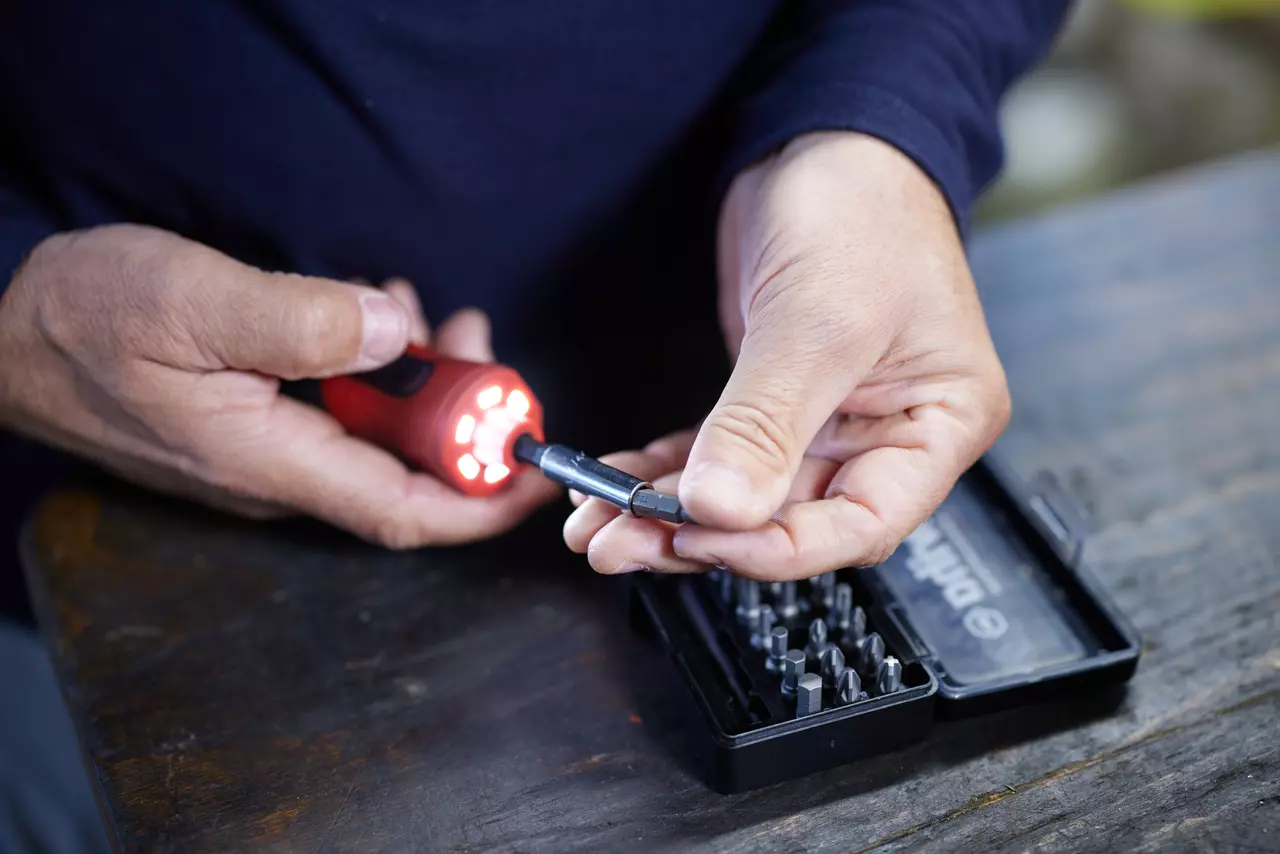 Person using an electric screwdriver with a bit set on a wooden table.