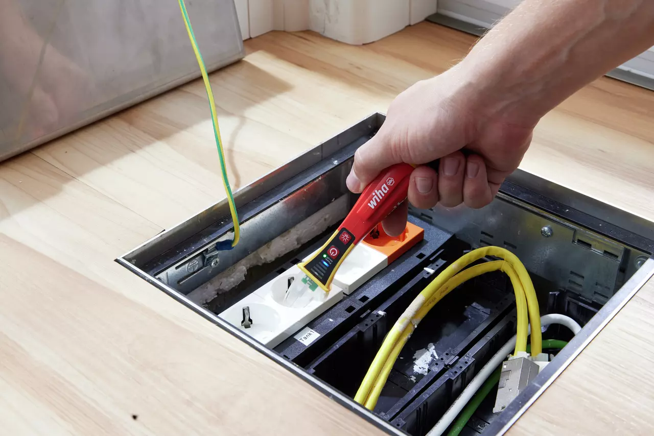 A person using a red non-contact voltage detector near a power socket in a floor box.