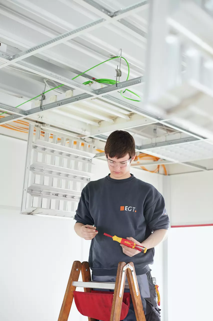 A person on a ladder working with an electric screwdriver near a ceiling with exposed wiring.