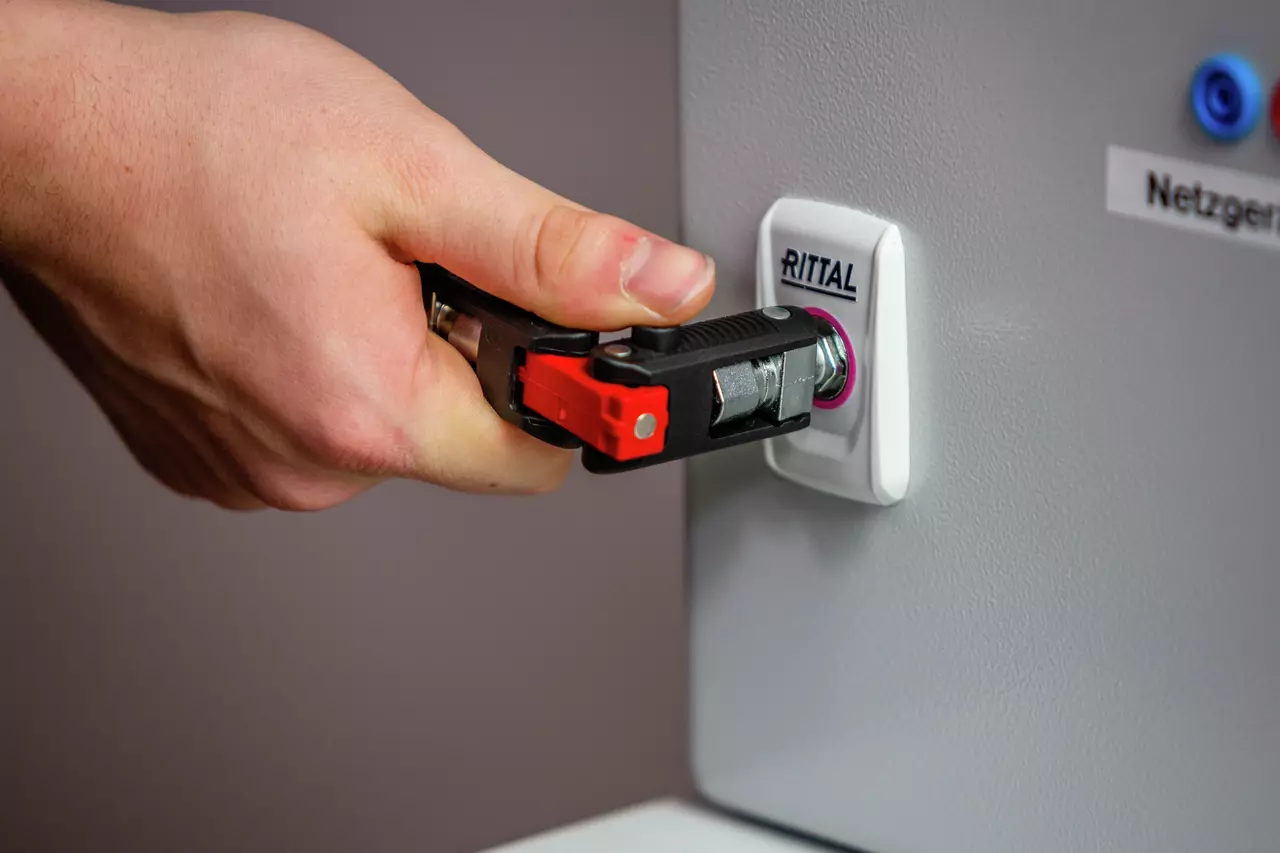 A hand using a black and red universal key on a lock labeled "Rittal" on a gray panel.