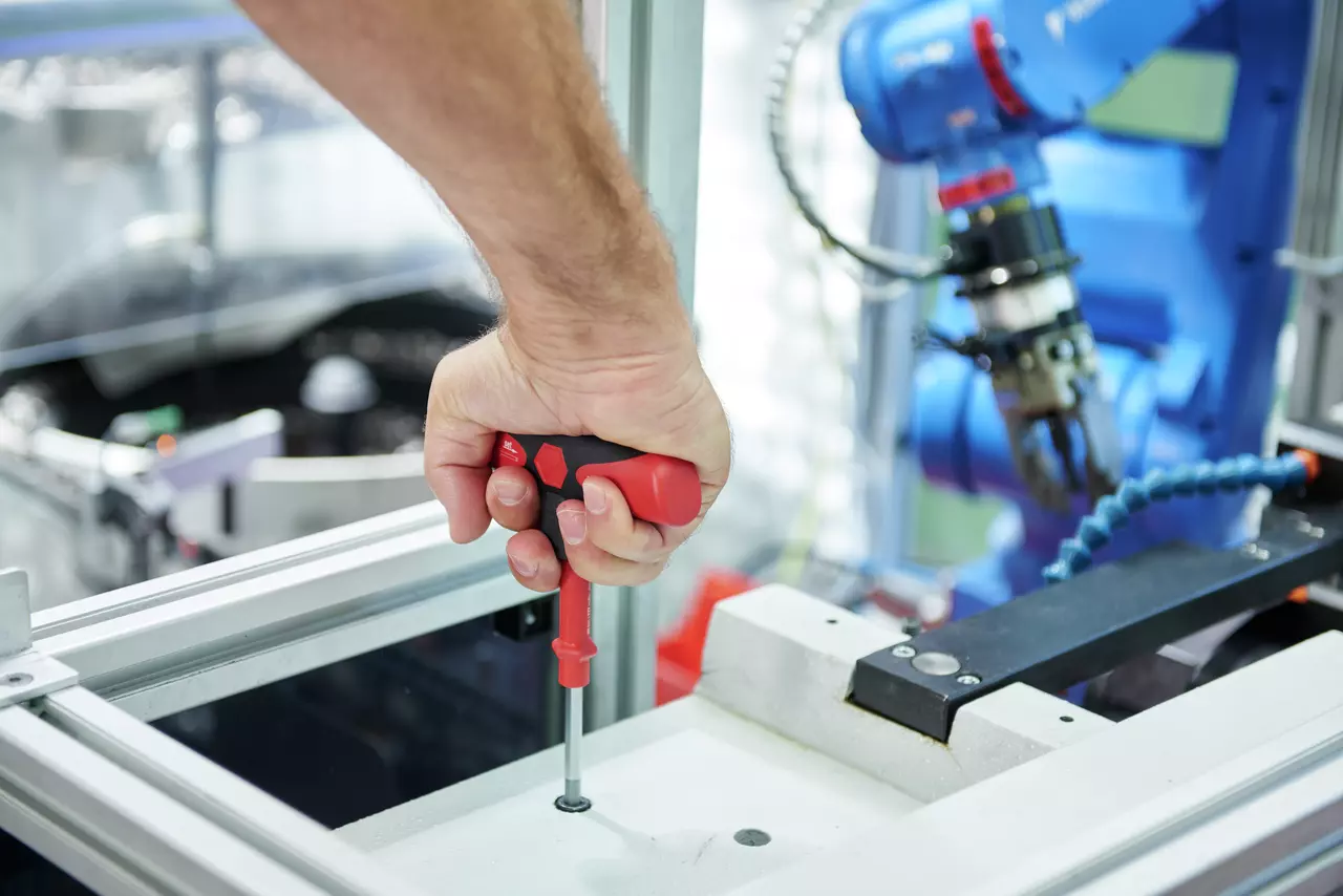 A person using a red and black torque screwdriver on a screw in a metal frame with a robotic arm nearby.