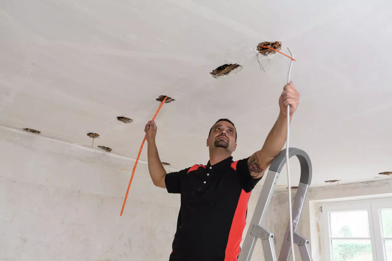 A person works on a ceiling, holding a measuring tape and a cable