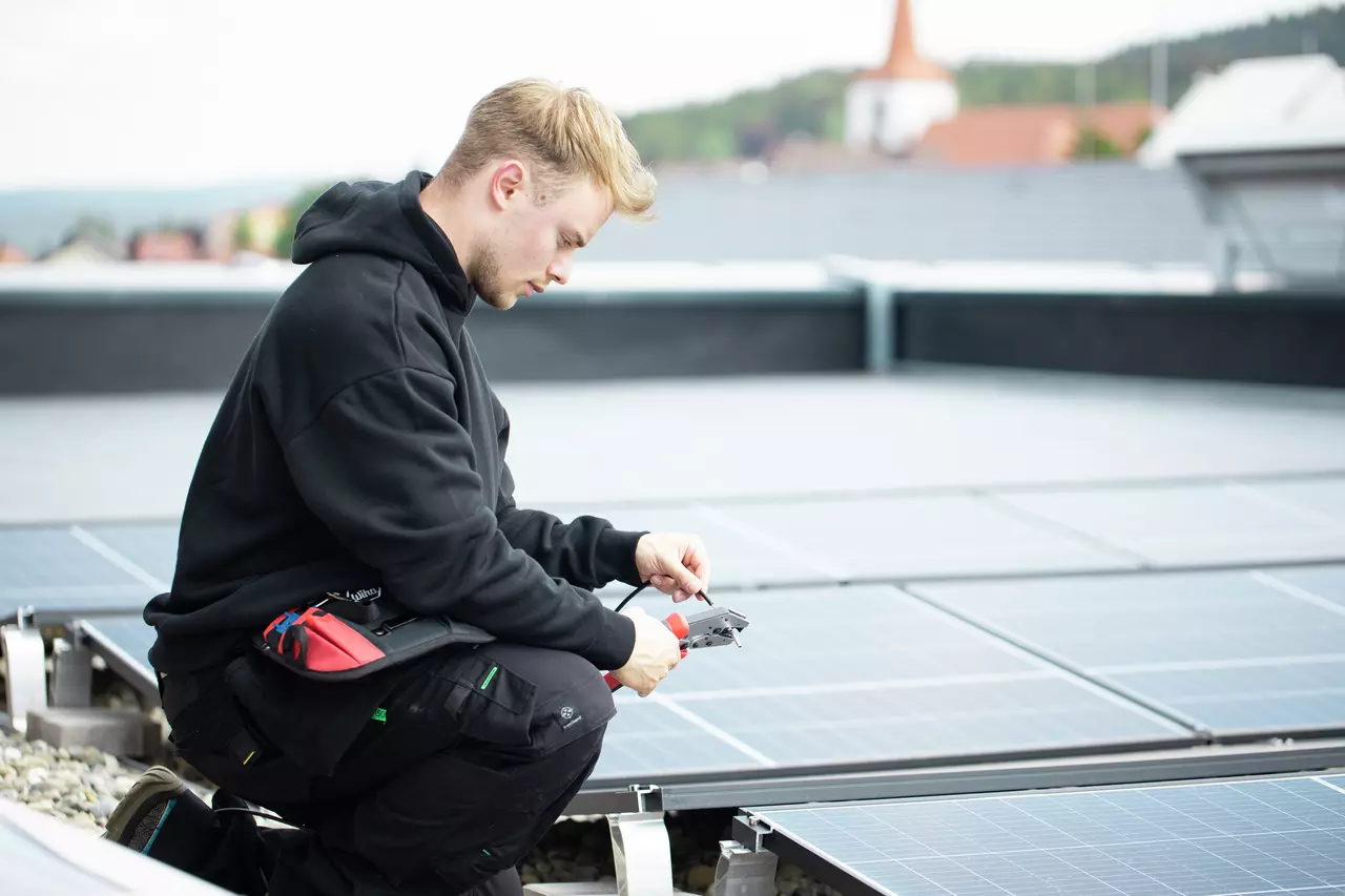 En person knæler på et tag og arbejder med et værktøj nær solpaneler.