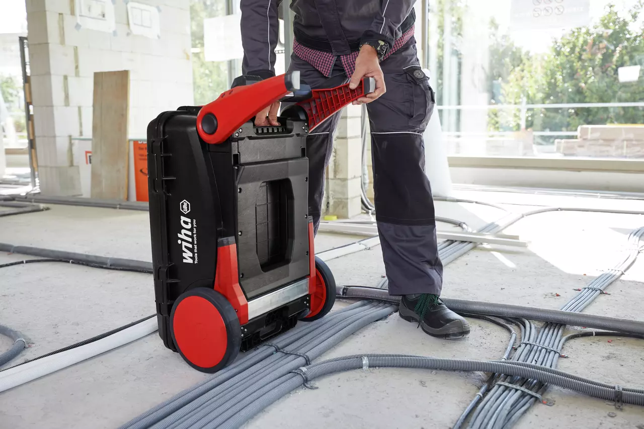 Person moving a large black and red tool case on wheels in a construction site.