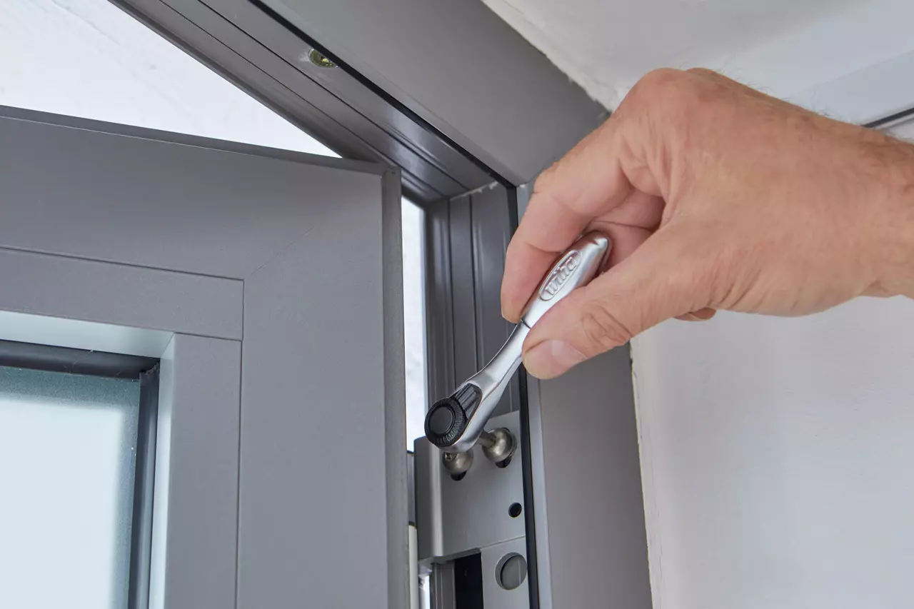 A person uses a ratchet to tighten a screw on a gray window frame.