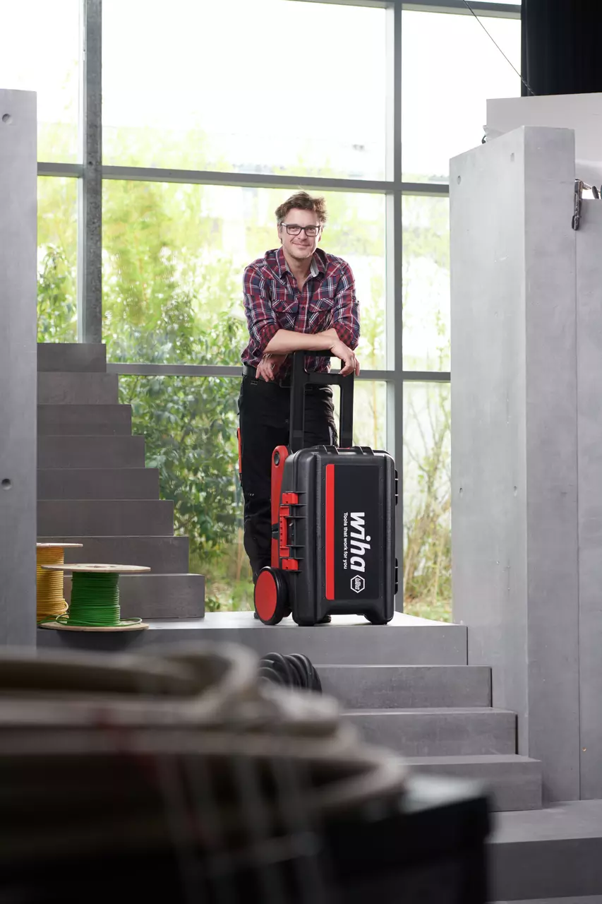 A person with glasses and checkered shirt stands on stairs with a large tool case with wheels.