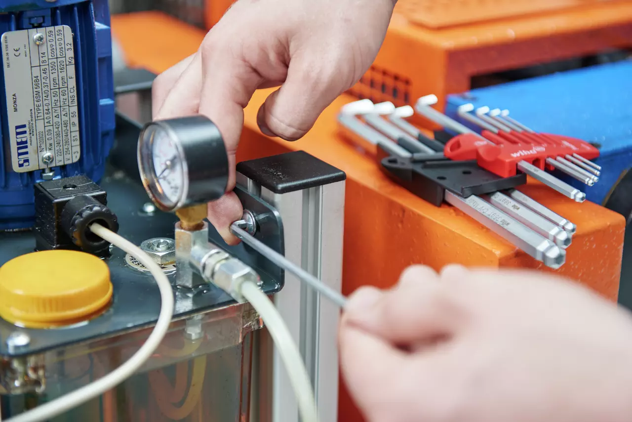 A person uses a hex key on a machine with a pressure gauge and cables nearby. Hex keys are on the side.