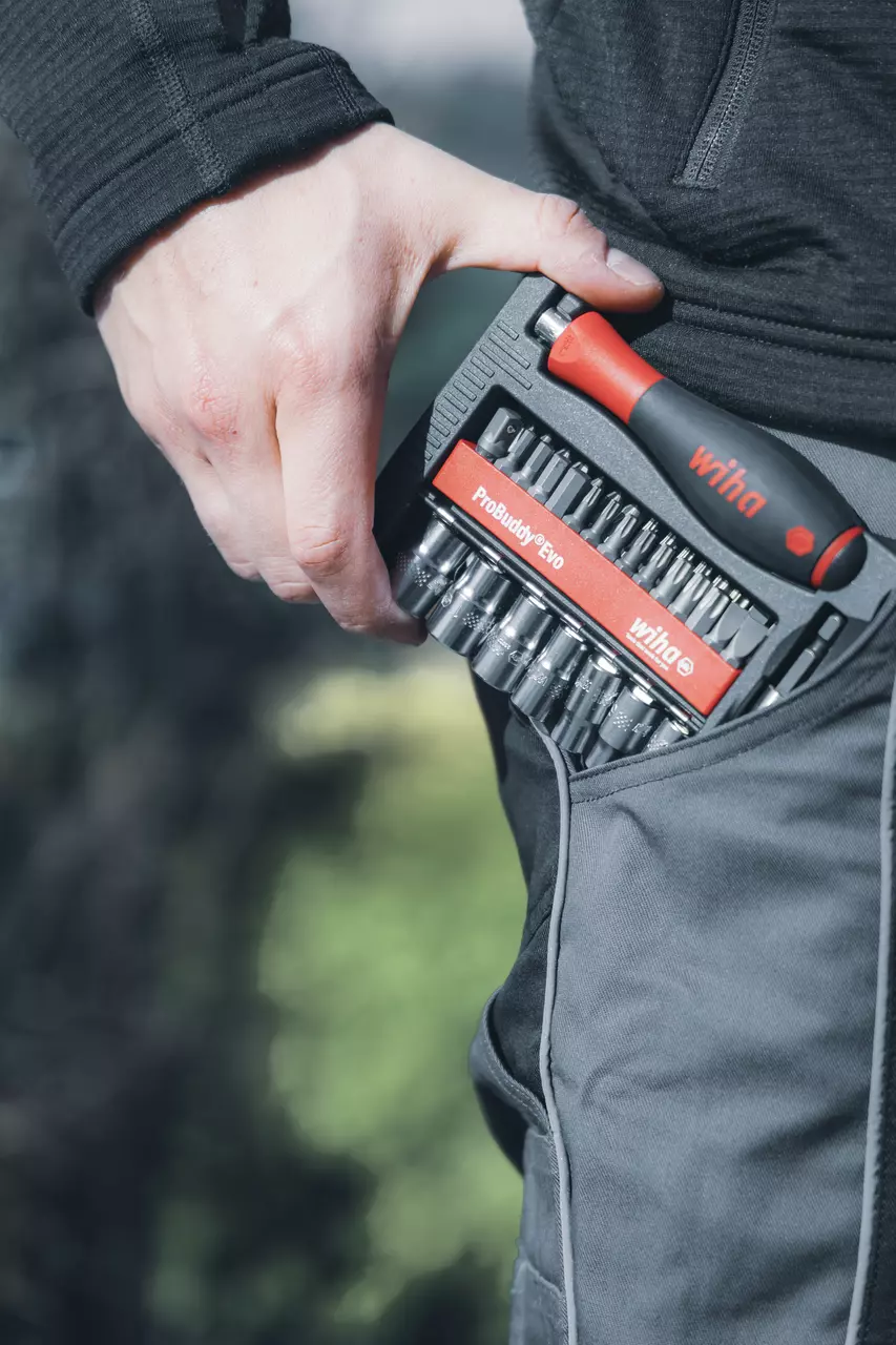 A person holds a tool set with a red and black handle and multiple bits in a trouser pocket.