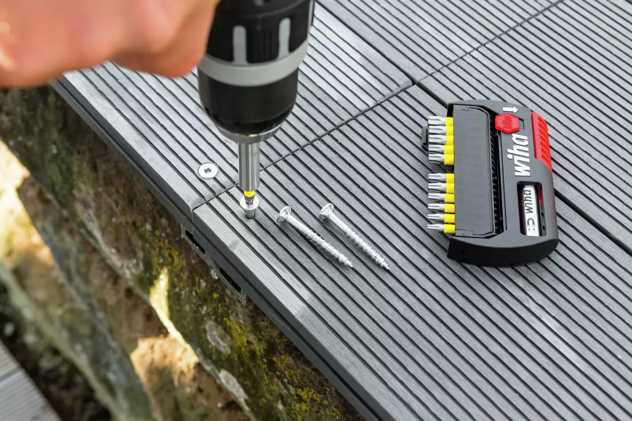 A person using a power drill to install screws on a grooved surface, with a bit set nearby.