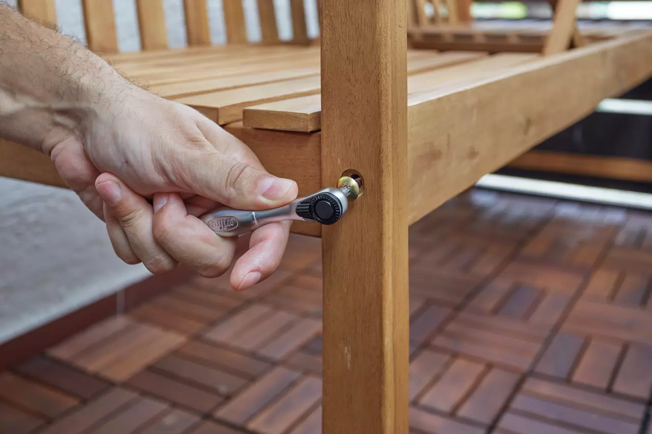 A hand using a ratchet wrench to tighten a bolt on a wooden bench.