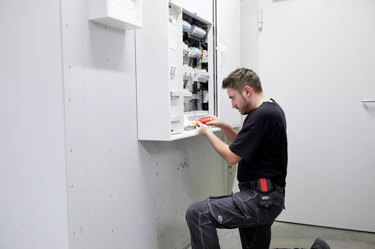 A person kneels while using an electric screwdriver on an open electrical panel