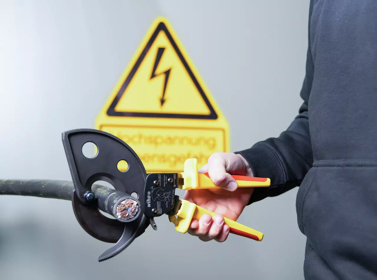 A person uses a ratchet cable cutter on a thick cable, with a high voltage warning sign in the background.