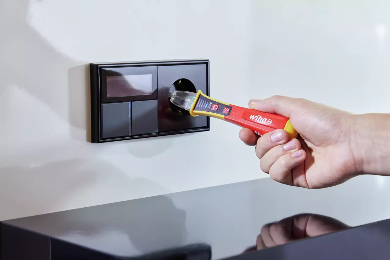 Hand using a non-contact voltage detector near a black wall socket.