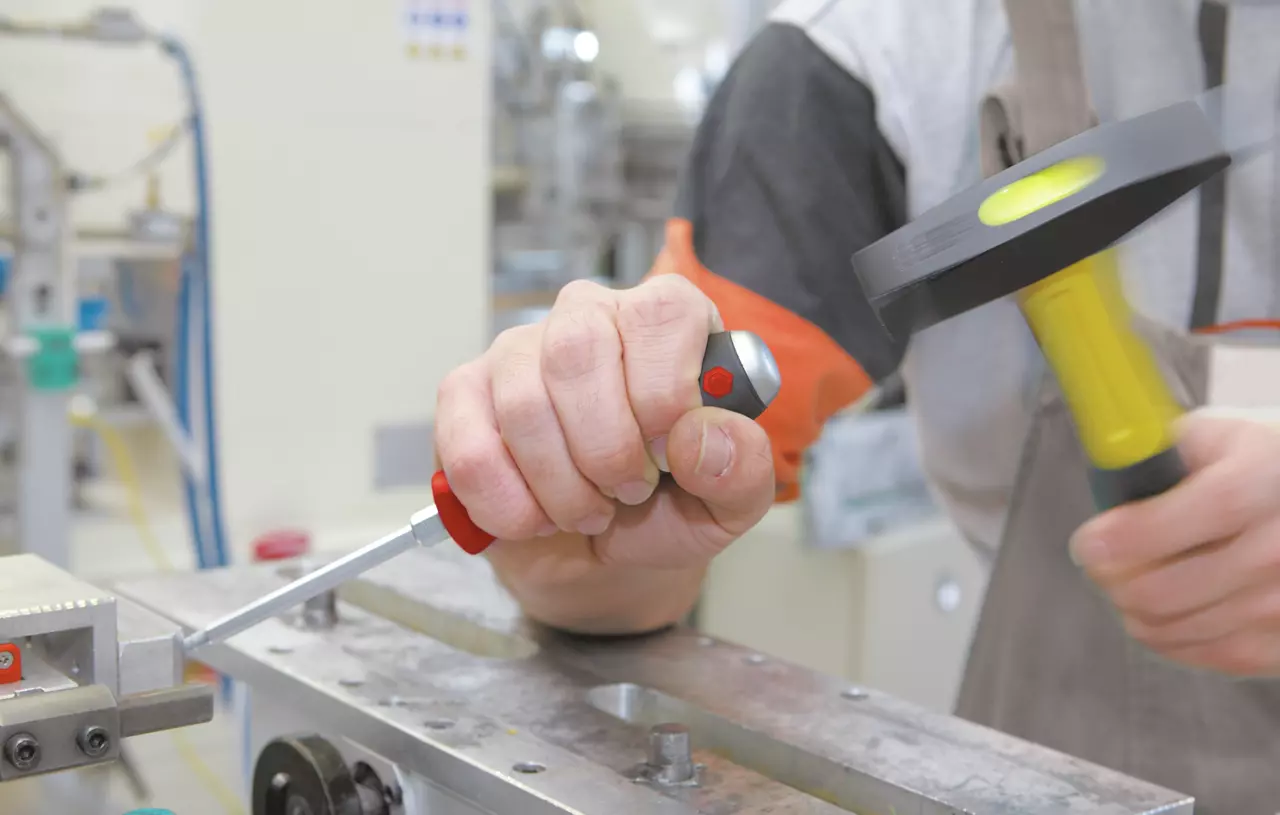 Person using a screwdriver with a metal cap and a hammer in a workshop setting.