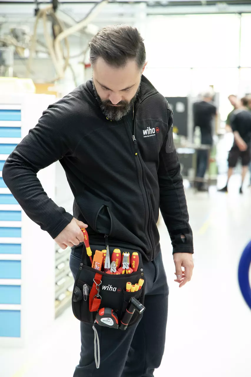 A person wearing a tool belt with various hand tools, including screwdrivers and pliers, in a workshop setting.