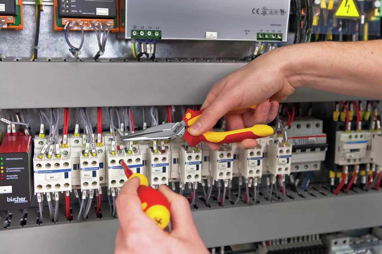 Two hands using pliers and screwdriver on electrical components in a panel.