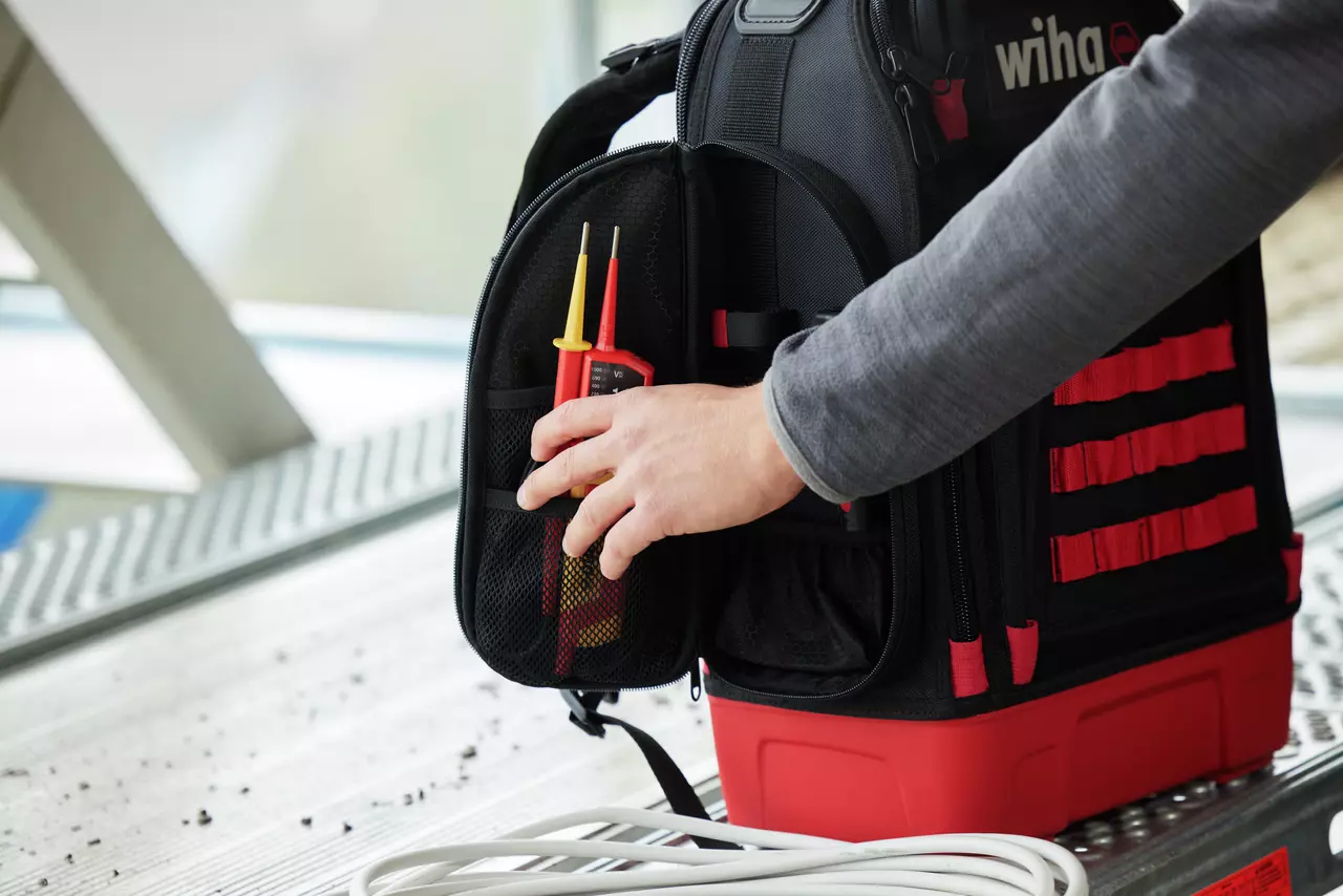 A person placing a voltage tester into a black and red tool backpack on a metal surface.