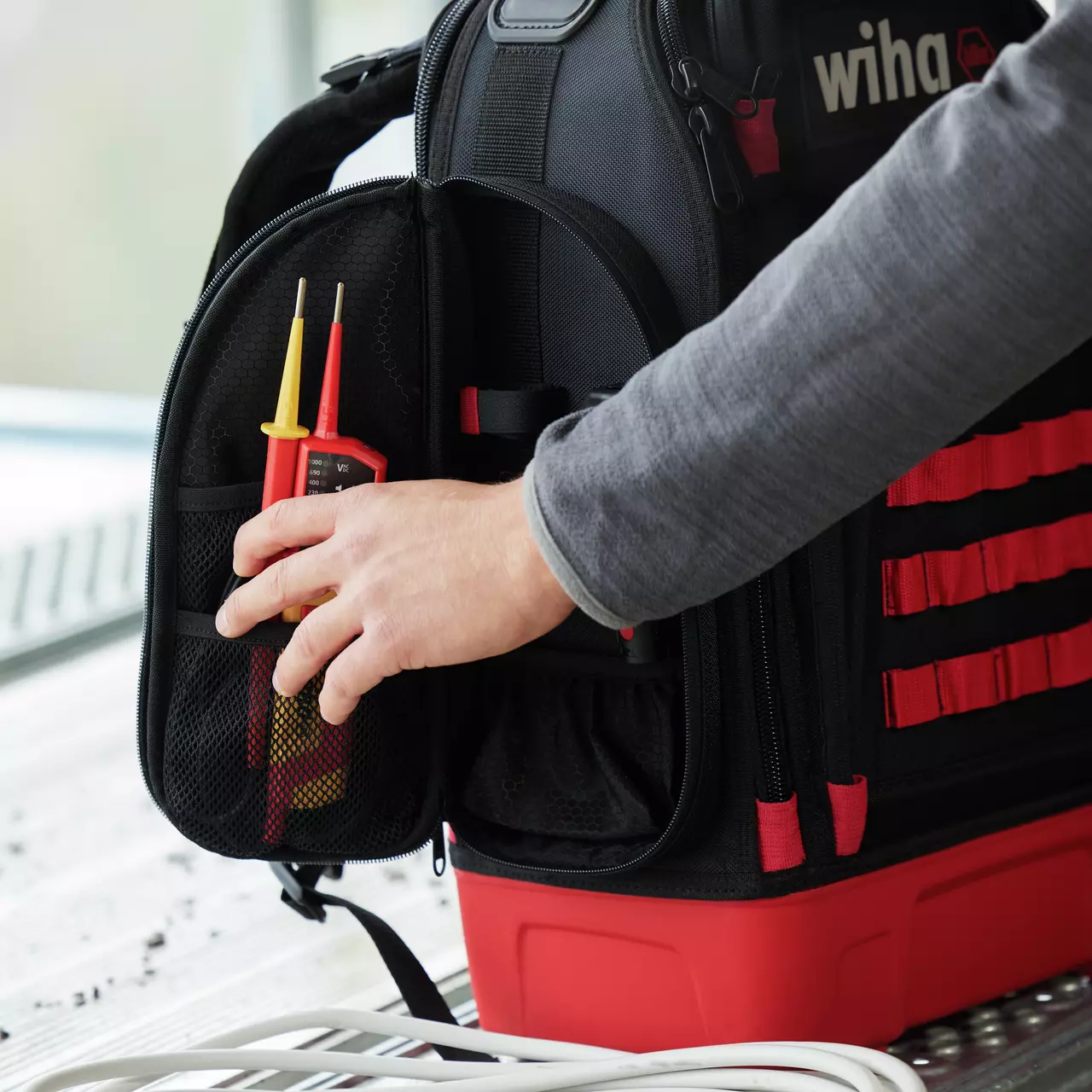 A person placing a voltage tester into a black and red tool backpack on a metal surface.