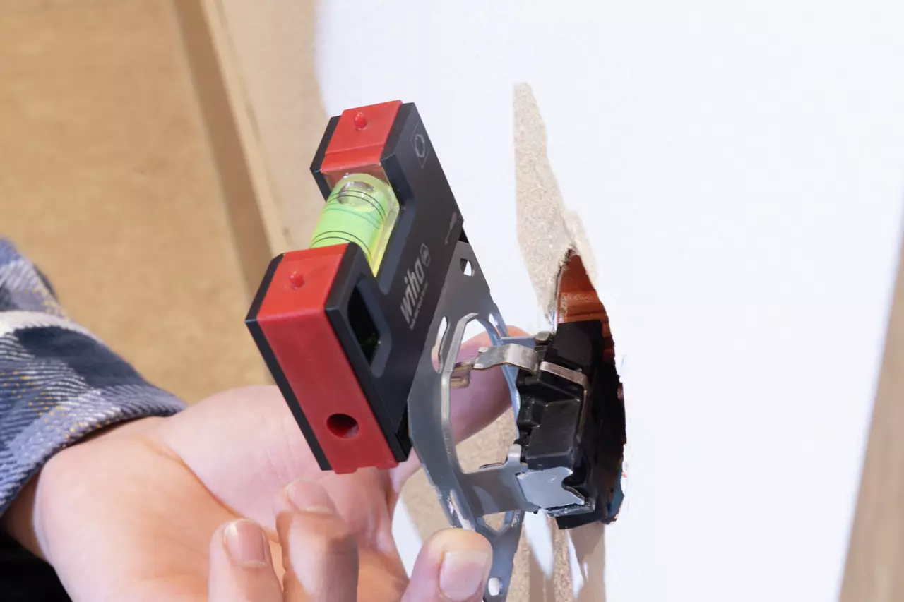 Person using a red and black pocket level to align a wall box in a cutout on a white wall.