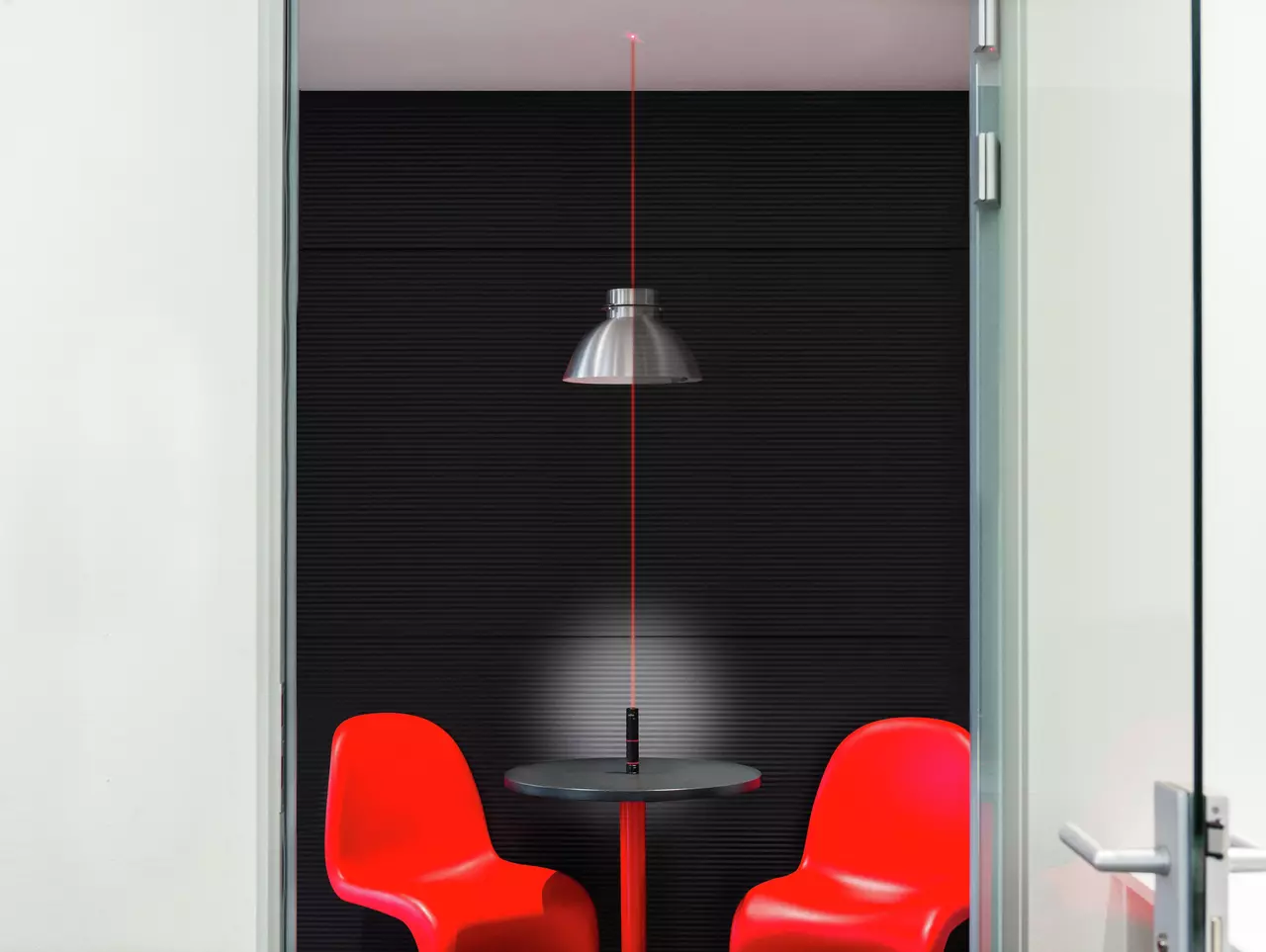 A red laser level projects a vertical line in a room with red chairs and a table under a ceiling lamp.