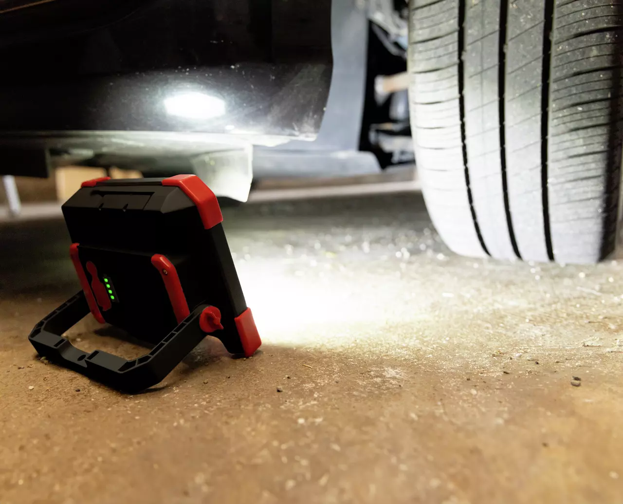 Portable work light illuminating under a car near a tire, placed on a concrete floor.