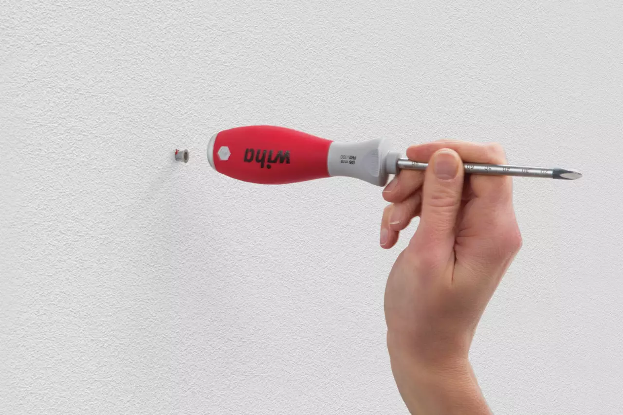 A hand uses a red-handled screwdriver to adjust a wall anchor on a white wall.