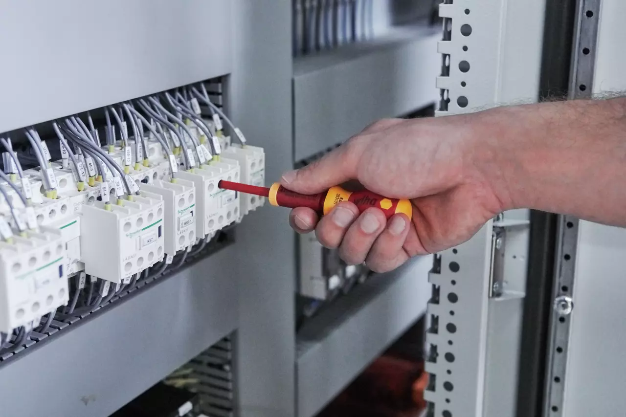 A person uses an insulated screwdriver on an electrical panel with multiple wires and switches.