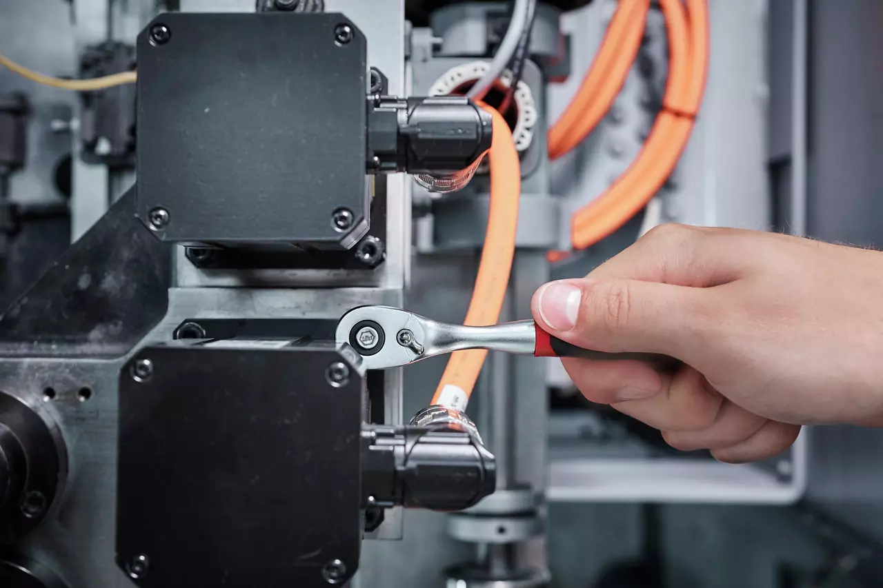 A hand using a reversible ratchet wrench on machinery with visible orange cables.