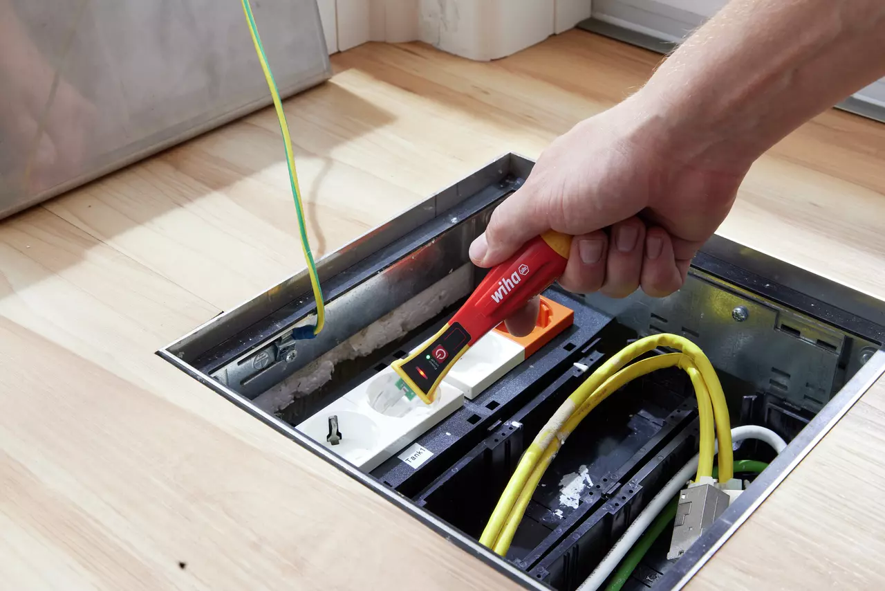 A person using a red non-contact voltage detector on a floor socket with cables