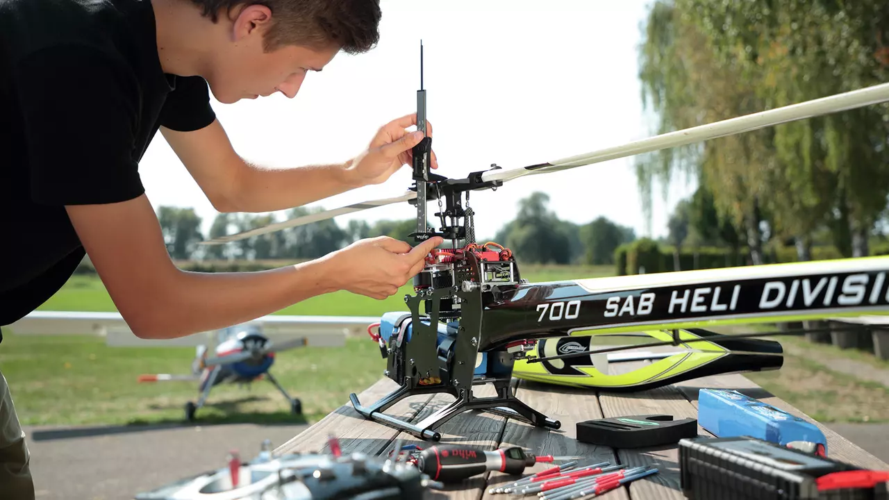 A person adjusts a model helicopter on a table outdoors, with tools scattered around.