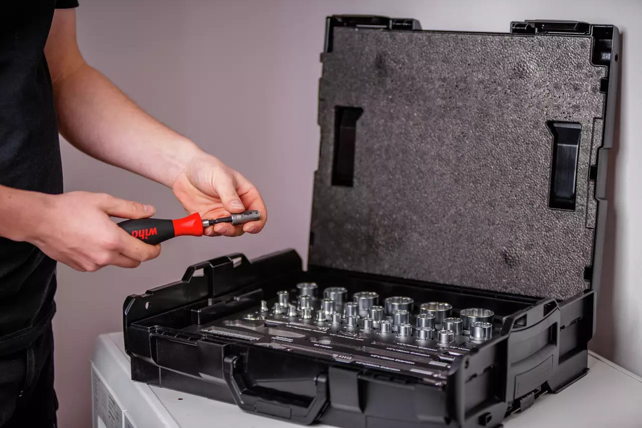 A person holding a tool with a red handle next to an open socket set case.