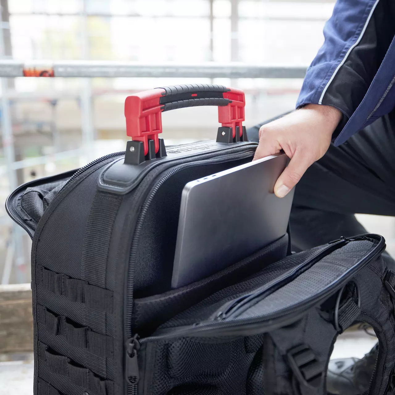 Person placing a laptop into a black backpack with red handles. The setting appears industrial.