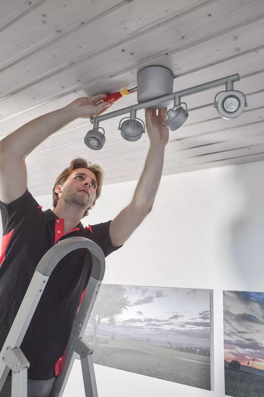 A person on a ladder installs a ceiling light using a screwdriver.