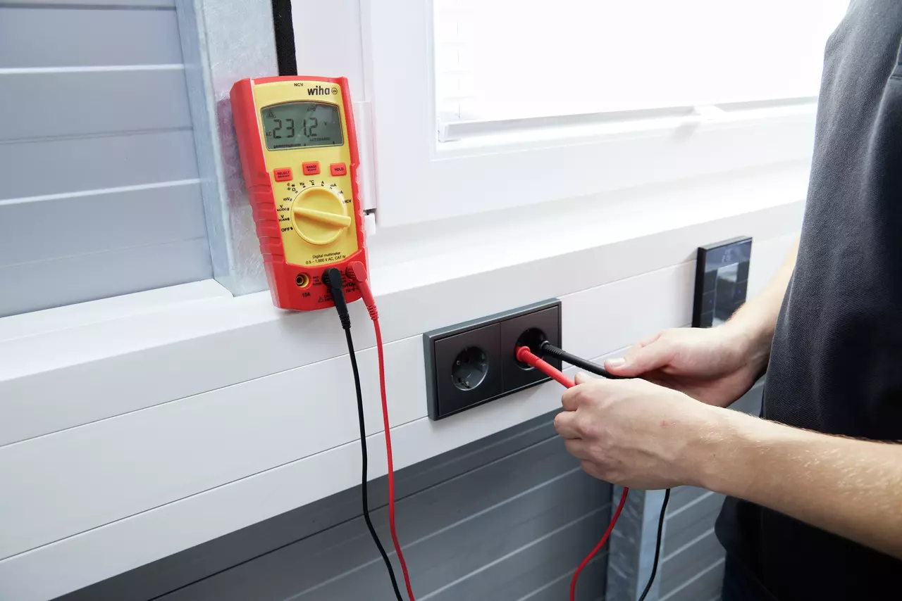 En person använder en digital multimeter för att mäta spänning i ett eluttag.