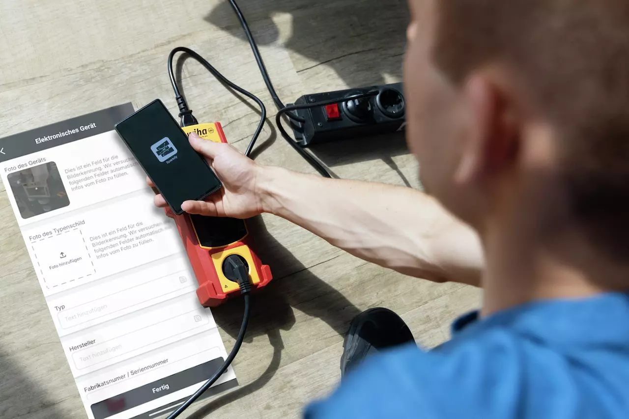 En person bruger en smartphone-app, mens de inspicerer en elektrisk enhed på gulvet.