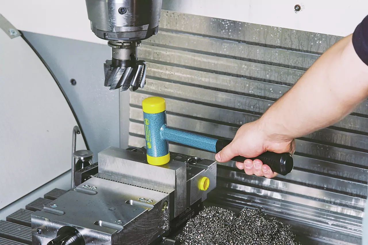 A person using a blue and yellow dead-blow hammer near a metalworking machine.