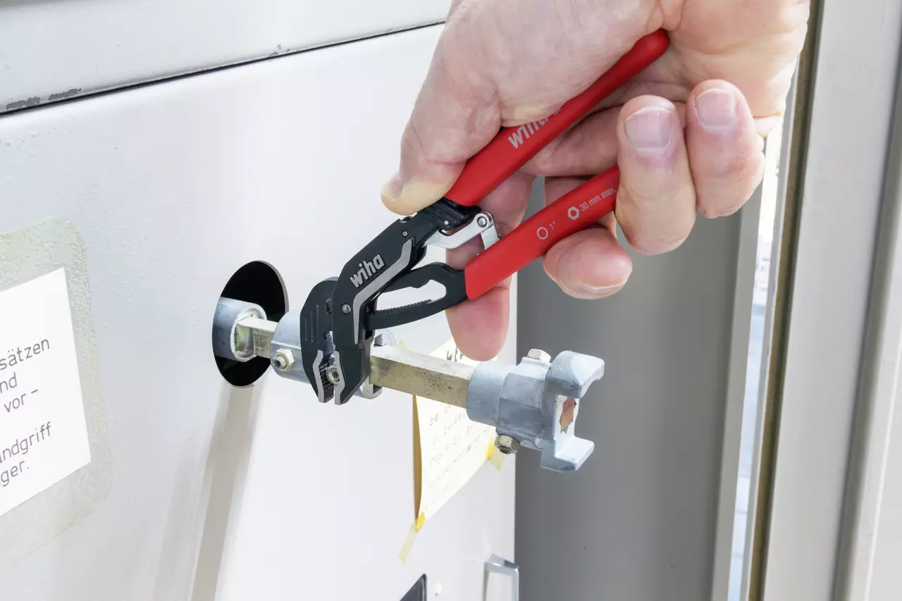 A hand holds a red water pump plier working on a metal pipe.