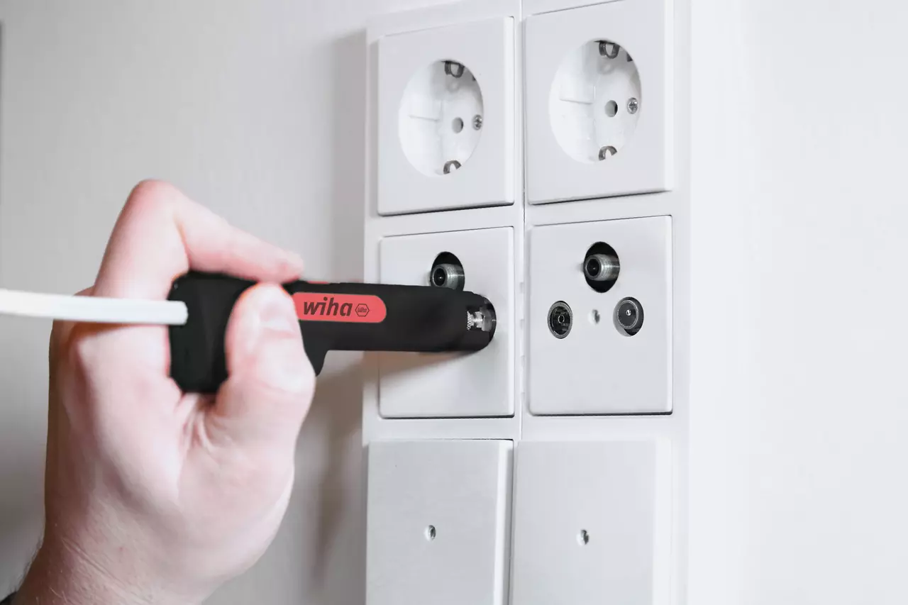 A person using a tool on a coaxial outlet panel. The tool has a black handle with a red logo.