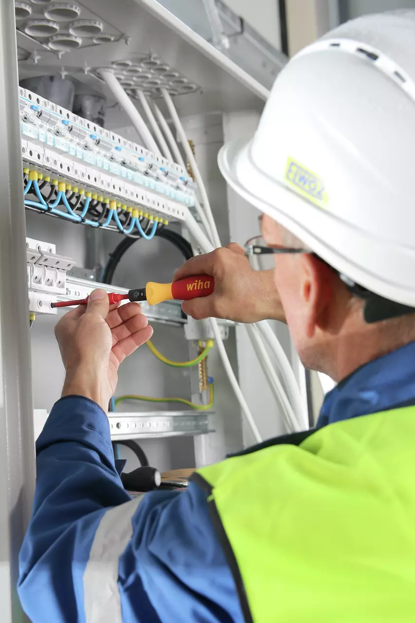 A person uses an electric screwdriver on an electrical panel, wearing a hard hat and safety vest.