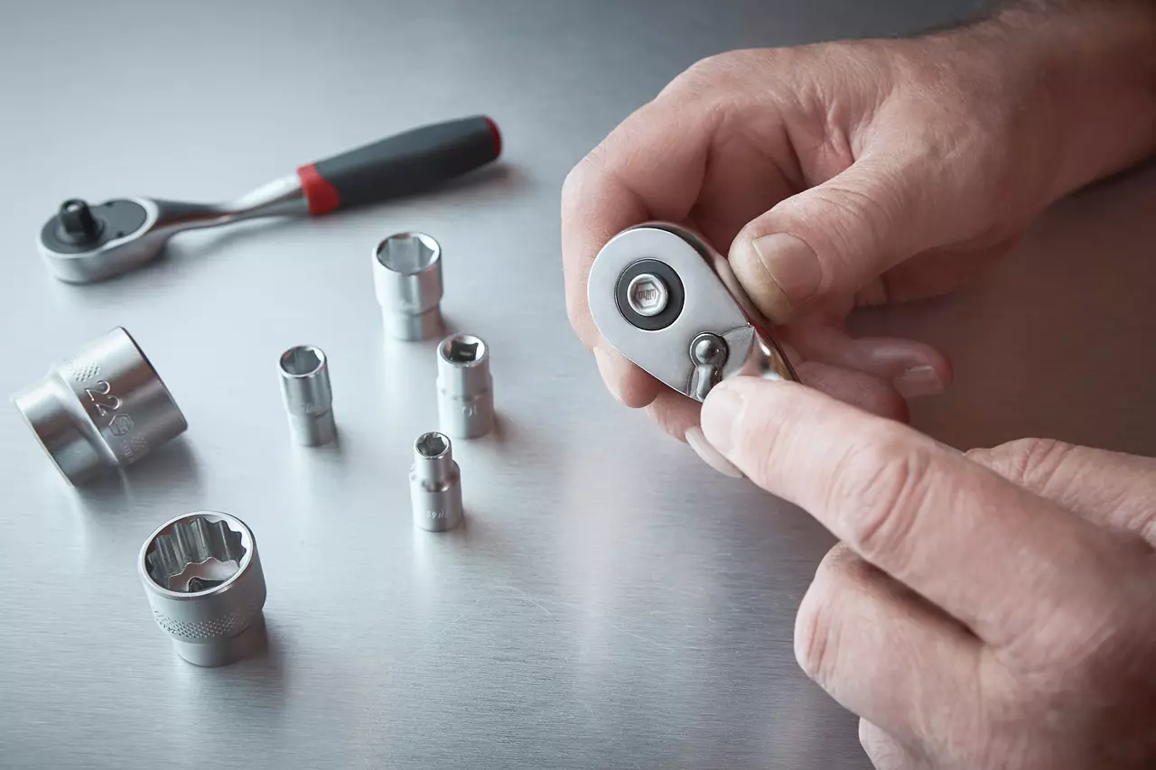 Hands holding a reversible ratchet, with various socket heads on a metal surface.