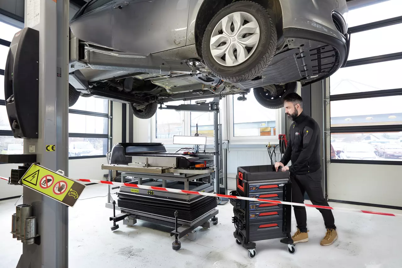A mechanic pushes a tool trolley in a garage with a car lifted on a hydraulic lift.