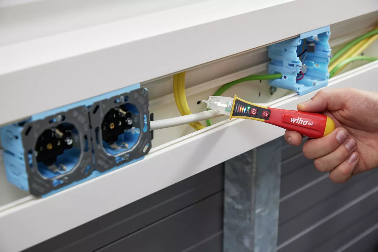Hand using a red non-contact voltage detector near electrical wires in a cable channel.